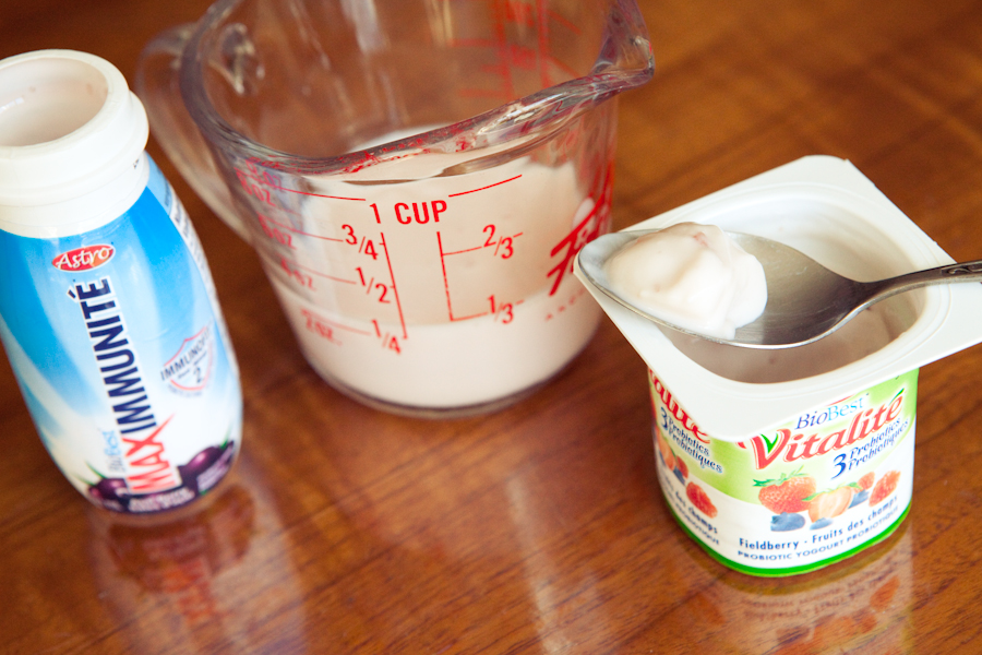 A photograph of a yogurt drink, measuring cup of milk, and a plastic cup of yogurt