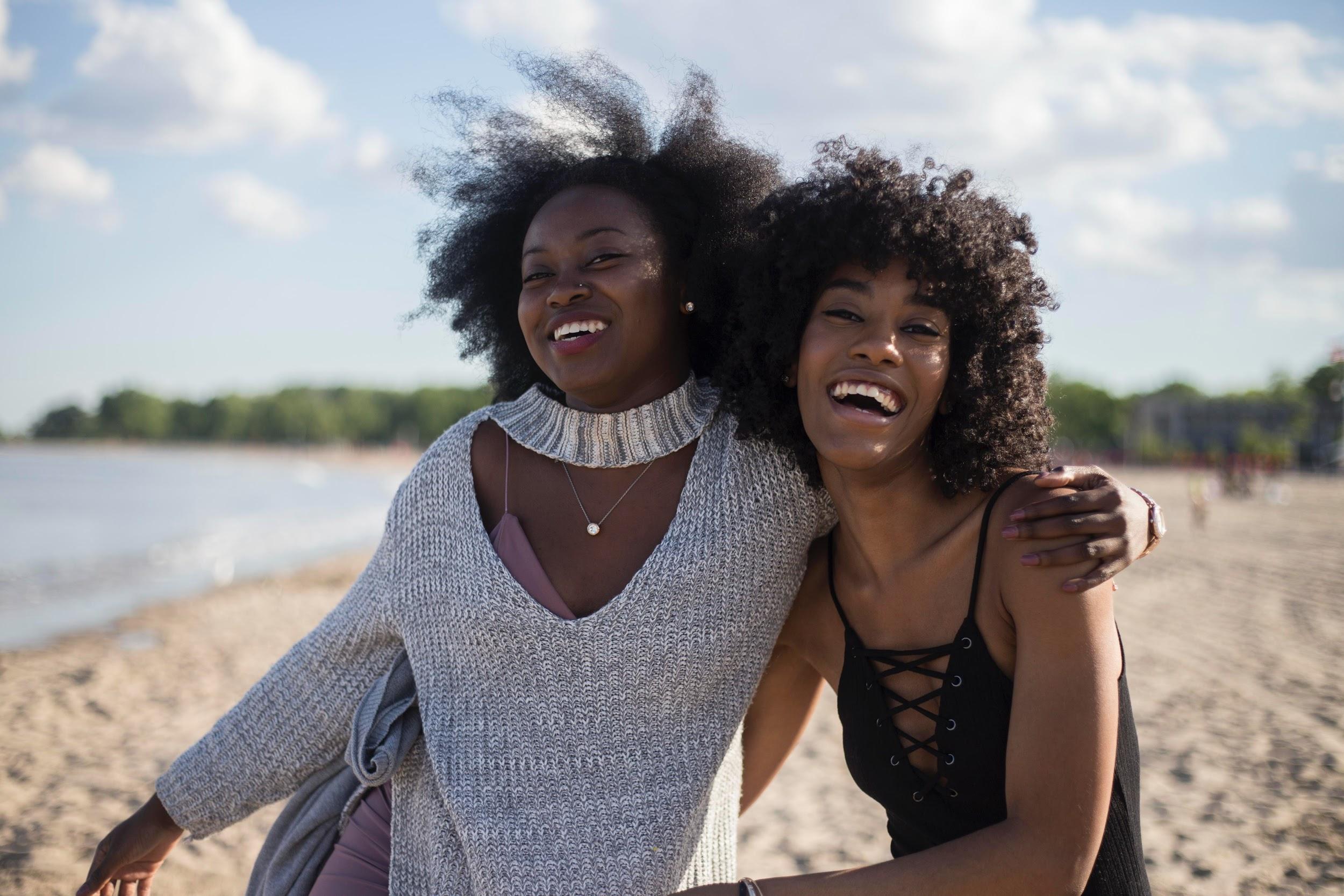 Two young black women smiling and hugging, while enjoying themselves on the beach.