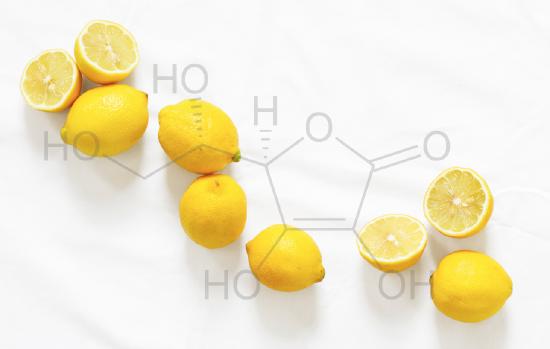 Several lemons are spread out on a white counter top. The chemical structure of vitamin C is shown superimposed over the lemons.