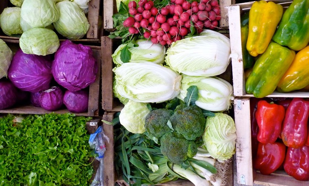 Photo shows a variety of colorful vegetable in bins, perhaps on display at a farmer's market. They include green and purple cabbage, red radishes, broccoli, leeks, lettuce, and red and yellow bell peppers.