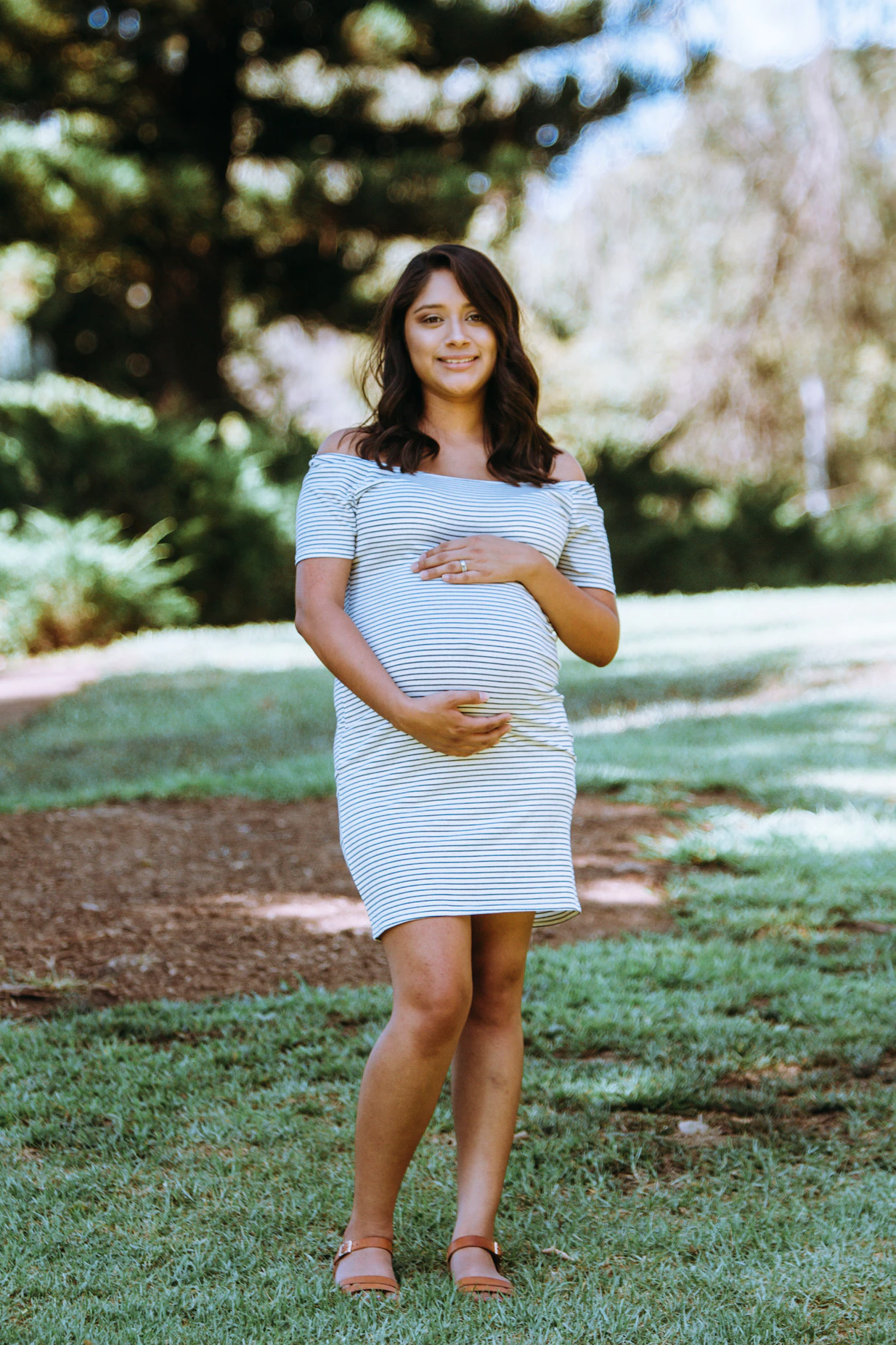 A pregnant woman stands outside with her arms encircling her belly