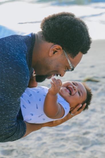 A Black father holds his baby on the beach. The father is smiling warmly, and the baby is laughing.
