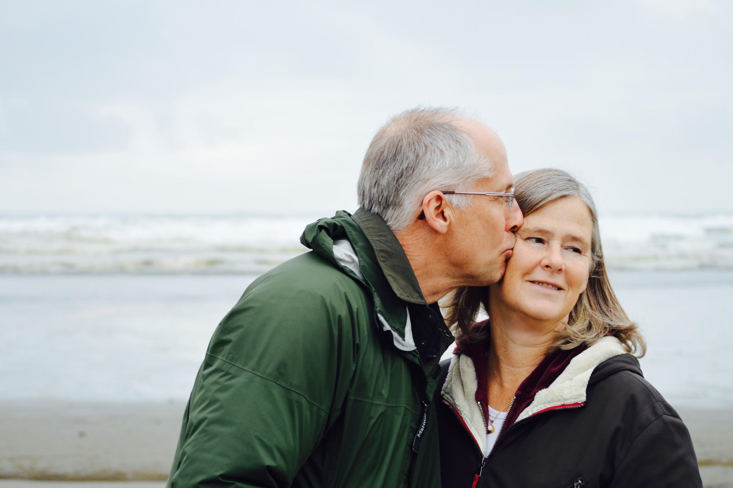 An older couple stand on the beach. The man is kissing the women on the cheek.
