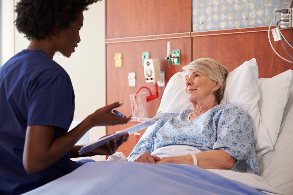 Imagen que muestra a una enfermera hablando con un paciente adulto mayor que se encuentra sentado en una cama de hospital