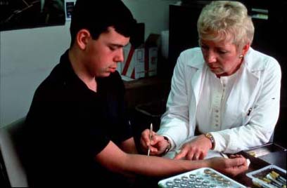 This image is of a young man receiving an allergy prick test. A blond-haired woman in a white lab coat sits on the right of the image. On her left is a young brown-haired man. His arm is out, and the woman, presumably a doctor, is pricking his skin with a tool. On the table in front of them is a tray of vials.