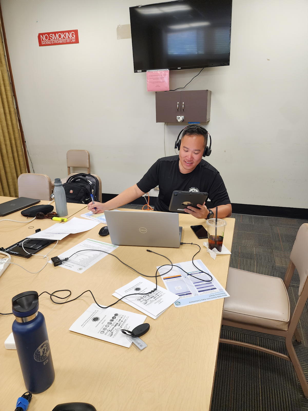Photo of a man wearing headphones sitting at a conference room table with papers, a laptop and other electronics spread out in front of him. 