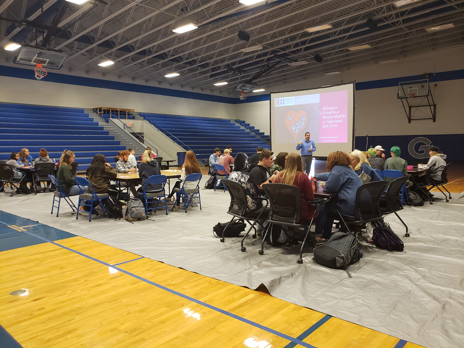 Photo of a person giving a presentation in a high school gymnasium, surrounded by people sitting at tables.