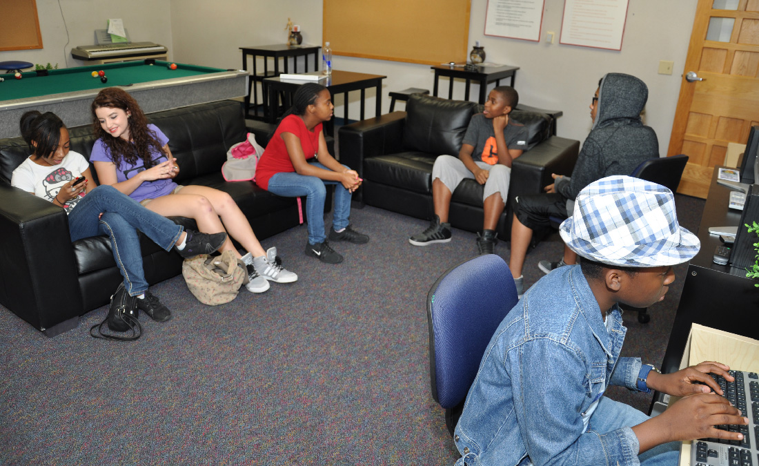 A color photograph shows several young boys and girls sitting on couches either talking or using computers.