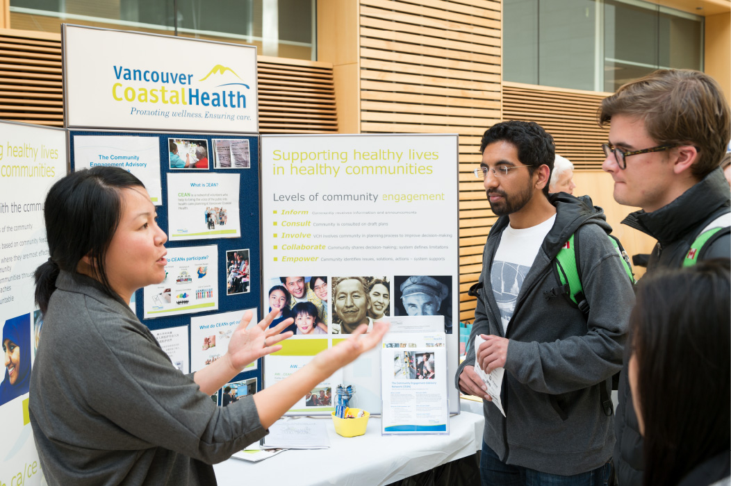 A nurse stands next to a table that holds a trifold posterboard and pamphlets with information from Vancouver Coastal Health. The nurse talks to two people standing in front of the table.