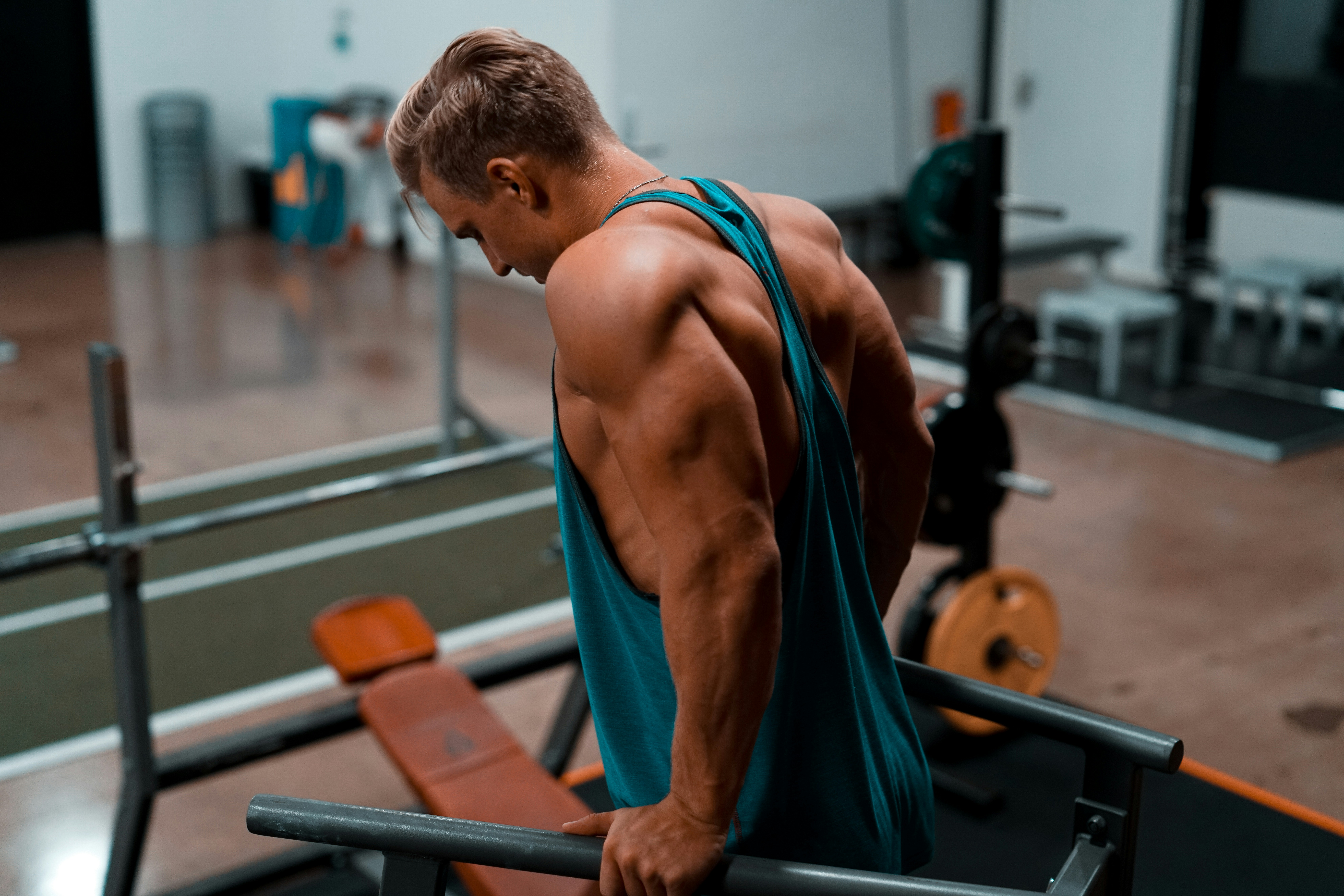 A muscular man in a tank top, works out at a dip station, which resembles parallel bars.