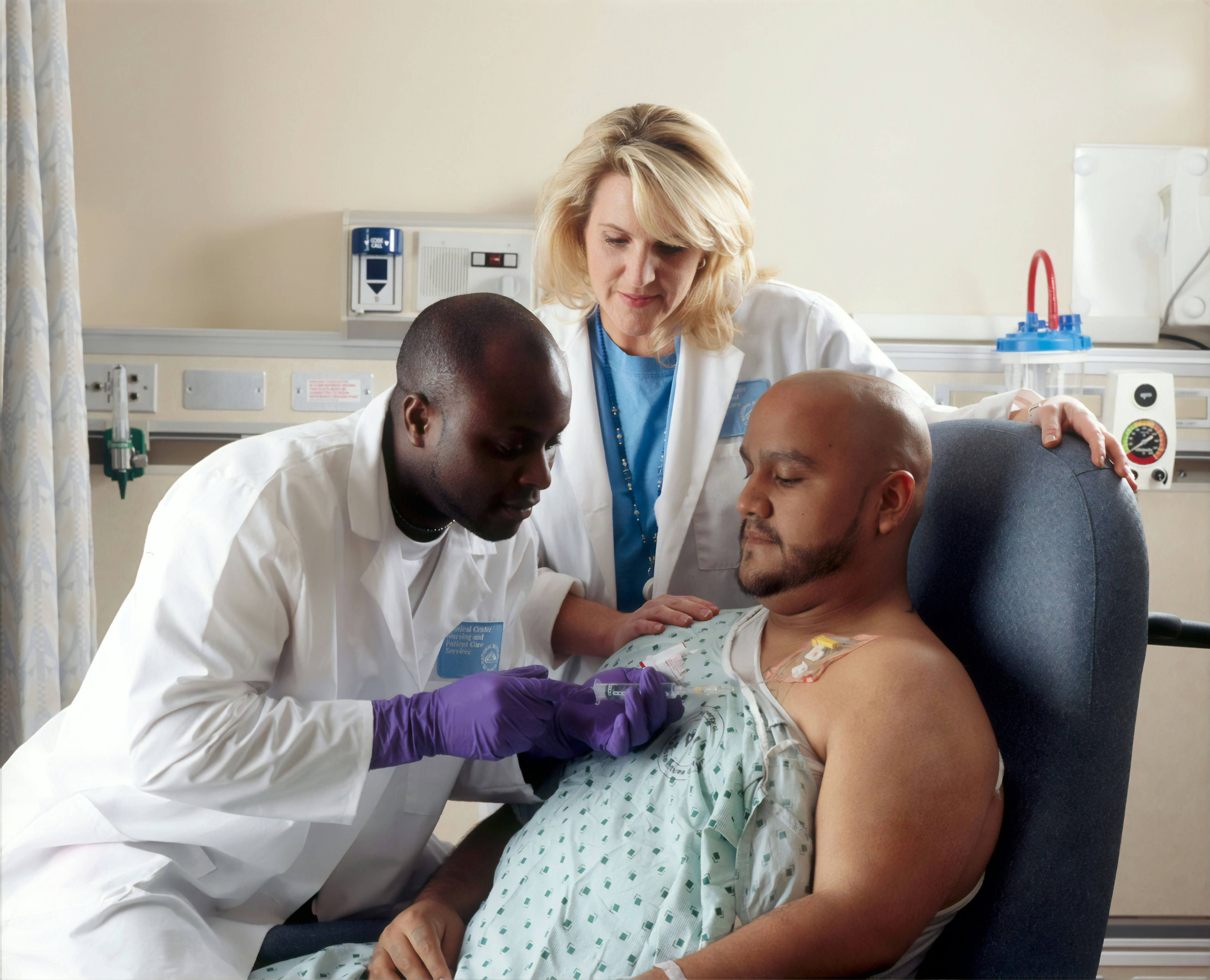 A man wearing a hospital gown watches as a doctor injects a medication into a tube that leads into his chest.