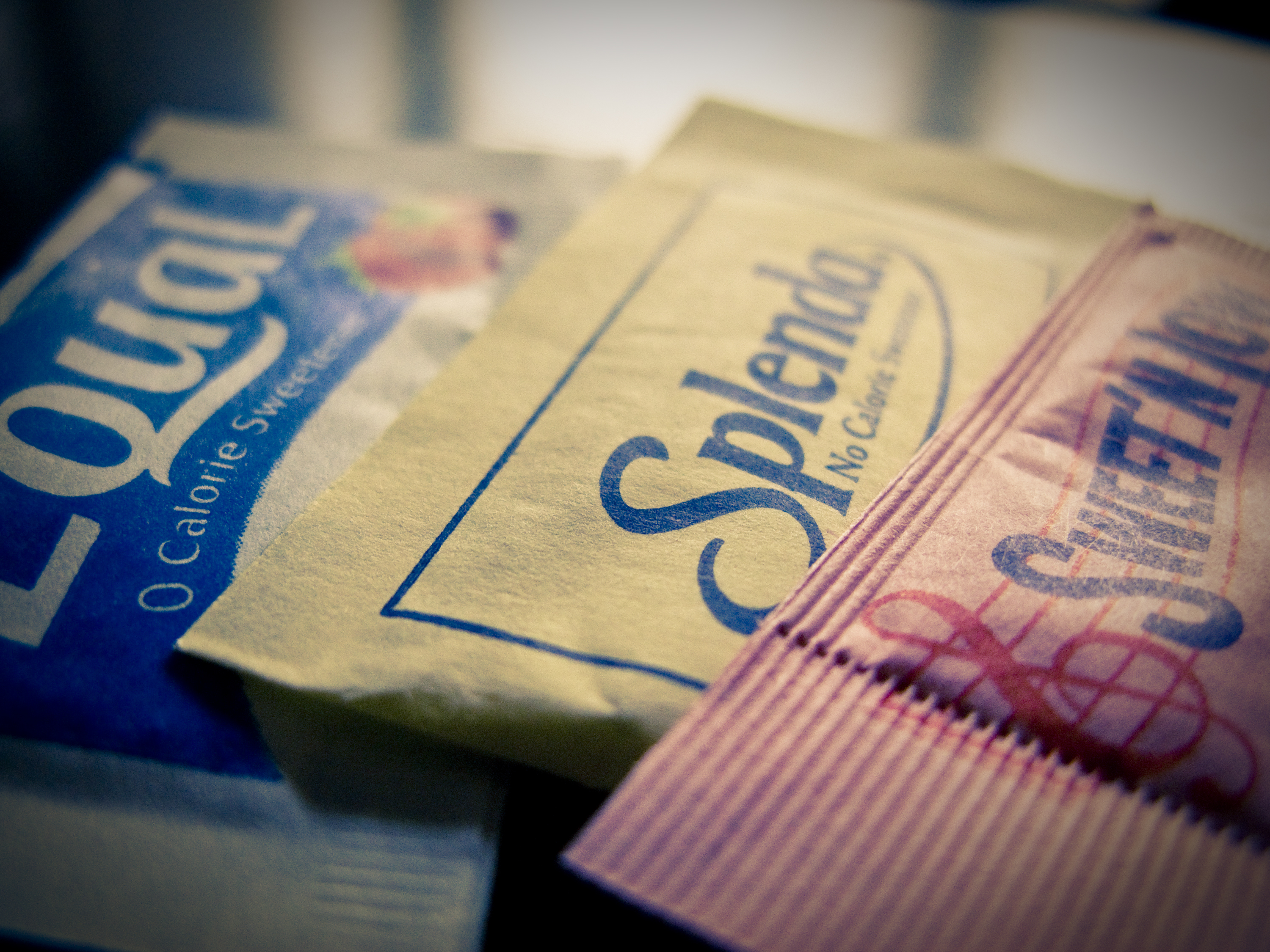 Three packets of artificial sweeteners: Equal, Splenda, and Sweet'N Low, stacked together on a dark surface.
