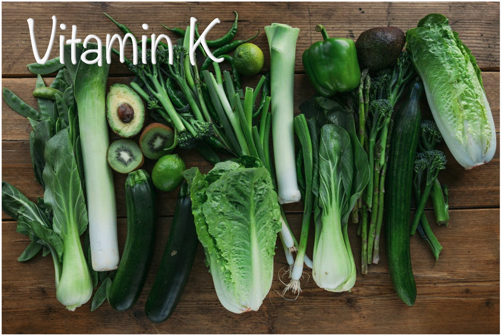 A variety of green vegetables and fruits, including avocado, zucchini, and broccoli, arranged on a wooden surface, labeled "Vitamin K."