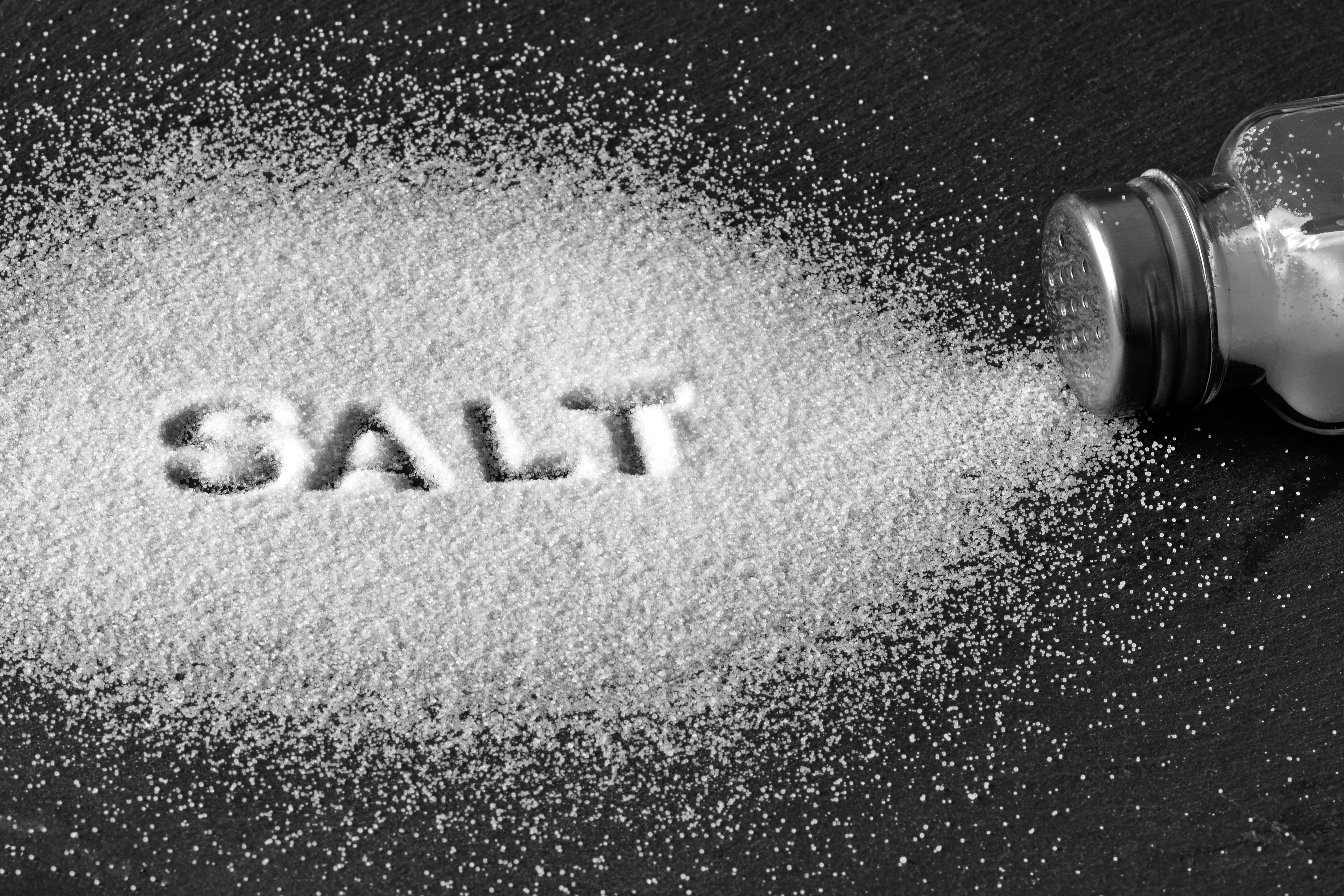The word "SALT" is formed in white salt on a dark surface, with a salt shaker spilling more salt nearby.