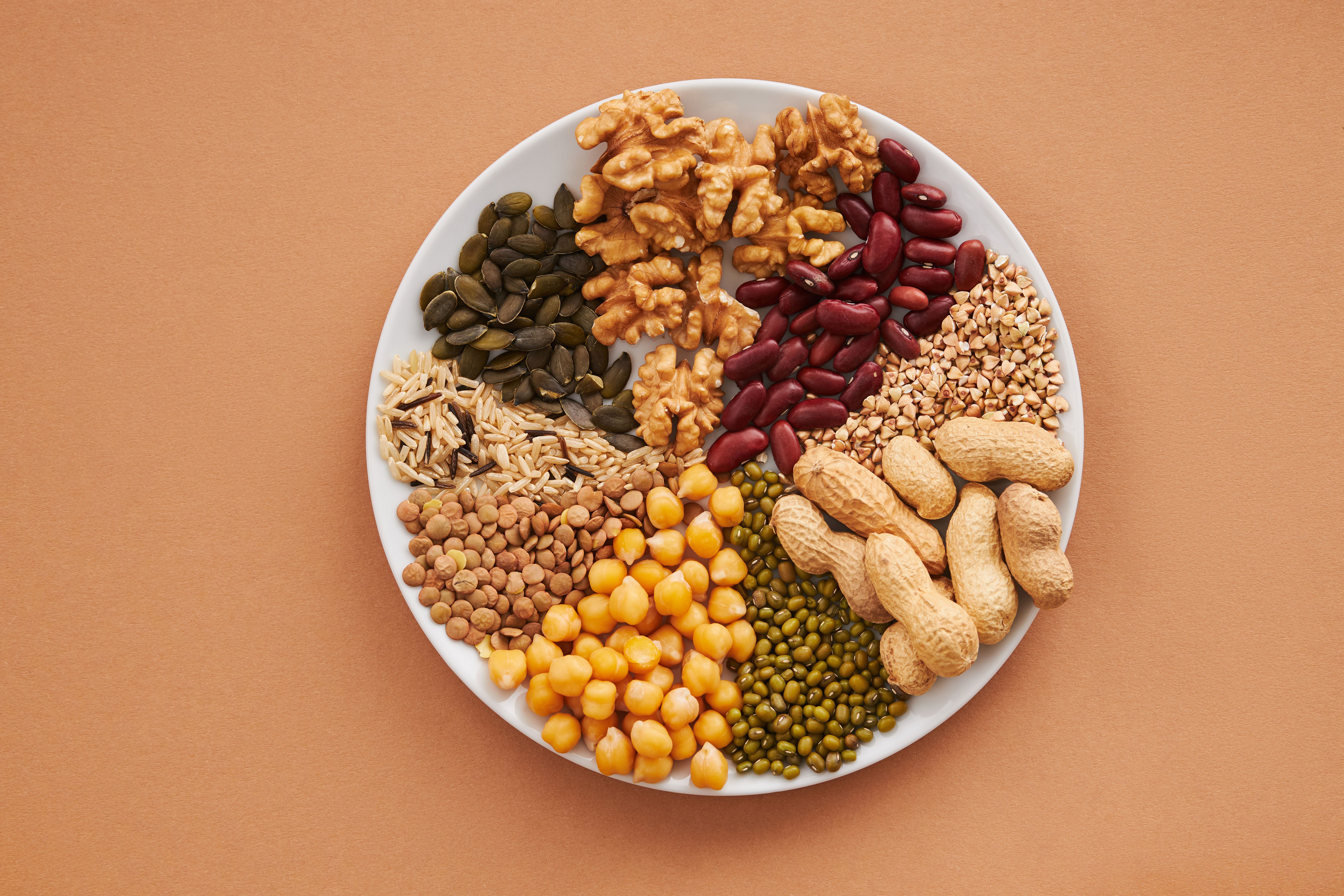 A round plate displays a variety of nuts and legumes, including walnuts, peanuts, chickpeas, lentils, and pumpkin seeds.